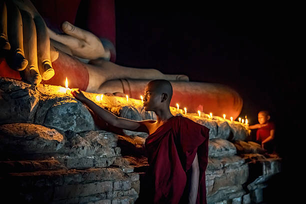 Two young Burmese buddhist monks in their typical clothing inside temple lighting up candles together to worship buddha. Natural Light, high ISO, inside dark temple with giant Buddha Statue. Bagan, Mandalay Region, Myanmar.